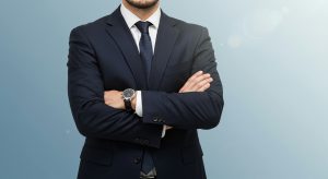 Postać ubrana w ciemny garnitur stoi z założonymi rękami na tle jasnego, jednolitego tła