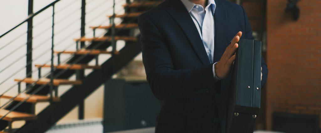 Na zdjęciu widzimy elegancko ubranego mężczyznę w granatowym garniturze i jasnej koszuli, trzymającego w dłoni czarną teczkę