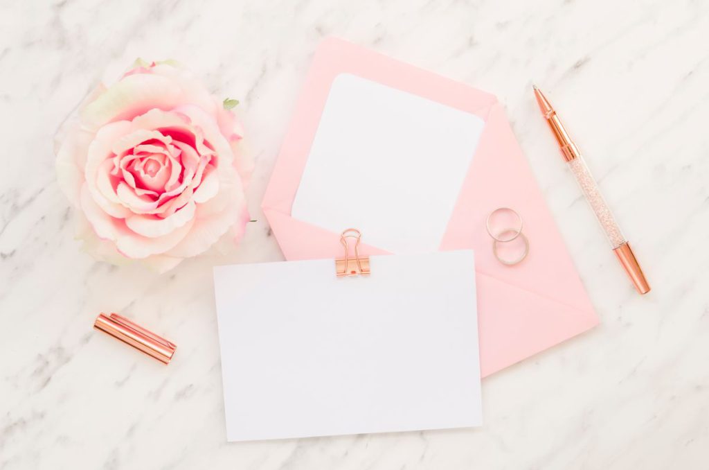 Eleganckie, czyste zaproszenia ślubne obok różowej koperty, obrączek i dekoracyjnej róży na marmurowym blacie.