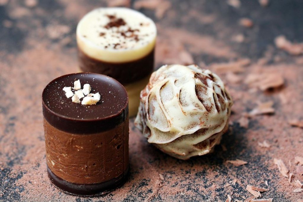 Trzy dekoracyjne praliny z czekolady deserowej i białej, stanowiące element eleganckiej bombonierki z logo, ułożone na ciemnym tle z posypką kakao.